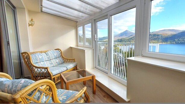 A balcony with wicker furniture, a glass table, and a view of a lake and mountains. - Otter Lodge 04 Loch Rannoch Highland Club (Pitlochry)