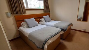 Ein Hotelzimmer mit zwei Betten, einem Fenster mit Vorhängen, einem Spiegel und einem hölzernen Kopfteil.