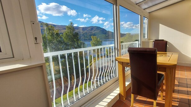 En balkon med et træbord og stole, udsigt til en sø og bjerge, samt et hvidt gelænder.