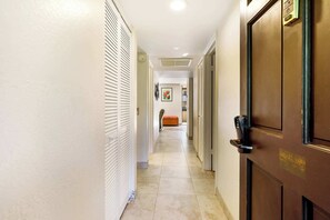 A hotel hallway with a wooden door, a white wall with a window, and a beige floor. - Honu Hale Minutes Away From the Beach! (Kailua-Kona)