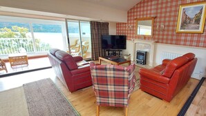 A living room with a red sofa, a plaid armchair, and a television. There is a fireplace and a large window overlooking a balcony. - Highland Lodge 43 Loch Rannoch Highland Club (Pitlochry)