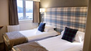 Une chambre d’hôtel avec deux lits, une fenêtre avec des rideaux, une tête de lit à motifs écossais et une lampe de chevet.