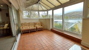 Une véranda avec un sol carrelé, deux fauteuils en osier et de grandes fenêtres offrant une vue sur des collines et un plan d’eau.