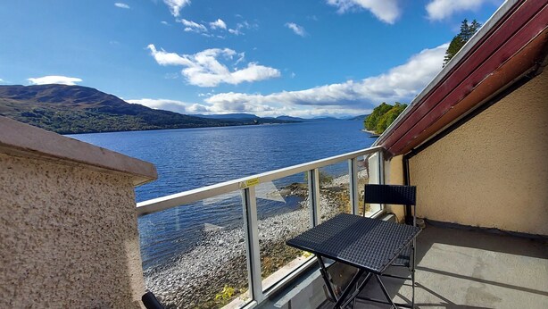 A balcony with a table and chair overlooking a lake and mountains. - Lochside Lodge 04 Loch Rannoch Highland Club (Pitlochry)