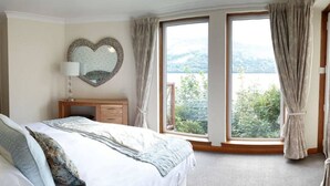 A bedroom with a large bed, a dresser, a heart-shaped mirror, and a view of greenery through a large window. - Lochside Lodge 04 Loch Rannoch Highland Club (Pitlochry)