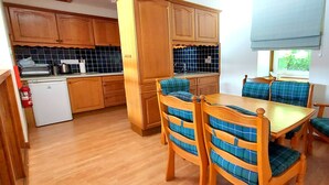 A compact kitchen with wooden cabinets, a white refrigerator, and a dining area with a wooden table and chairs with blue cushions. - Lochside Lodge 04 Loch Rannoch Highland Club (Pitlochry)