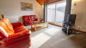 A living room with a red leather sofa, a wooden coffee table, a television on a stand, and a large window with a view of a body of water. - Lochside Lodge 04 Loch Rannoch Highland Club (Pitlochry)