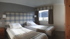 A hotel room with two beds, a large window with curtains, a headboard with a plaid pattern, and a nightstand with a lamp. - Osprey Lodge Loch Rannoch Highland Club (Pitlochry)