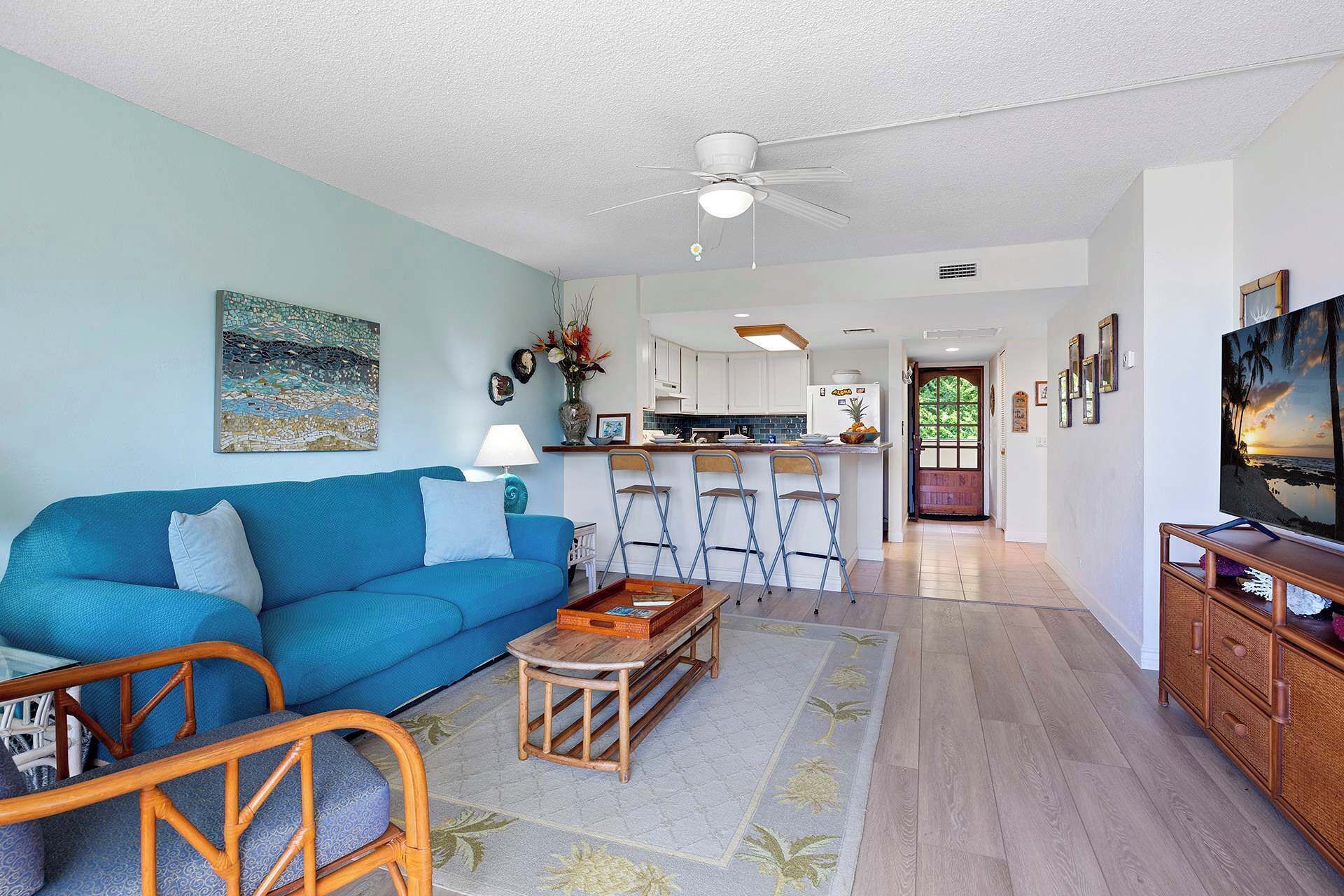 A living room with a blue sofa, a coffee table, and a TV on a wooden cabinet. There is a kitchen in the background with white cabinets and a dining area with bar stools.