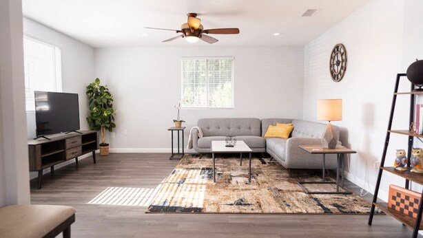 A modern living room with a grey sofa, a television, a ceiling fan, a window, and a decorative clock on the wall.