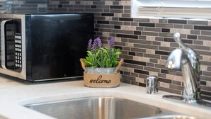 A kitchen with a stainless steel sink, a black microwave, and a decorative plant in a burlap pot with a 'welcome' sign.