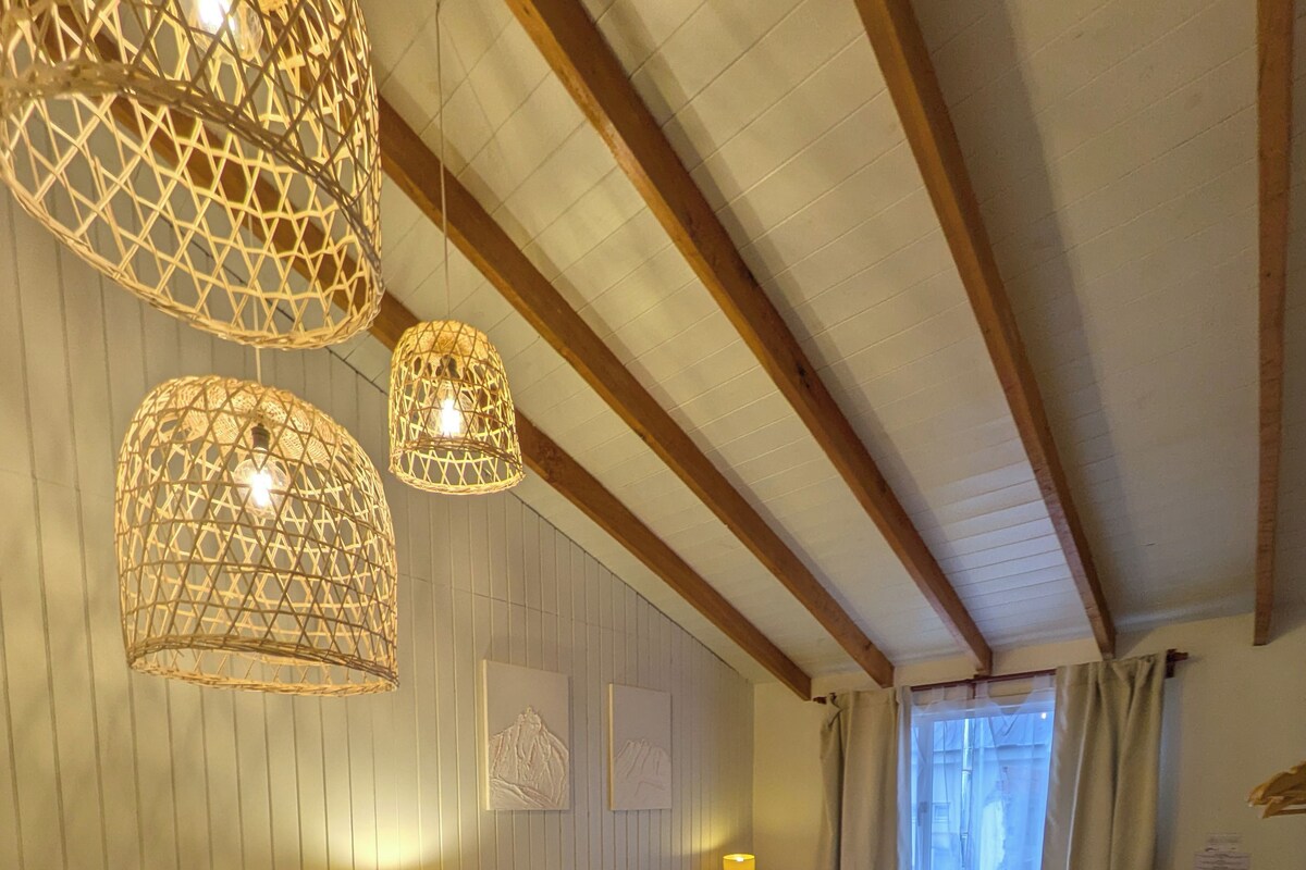 A room with a bed, two bedside lamps, a wicker chair, a small table, and a window with curtains.