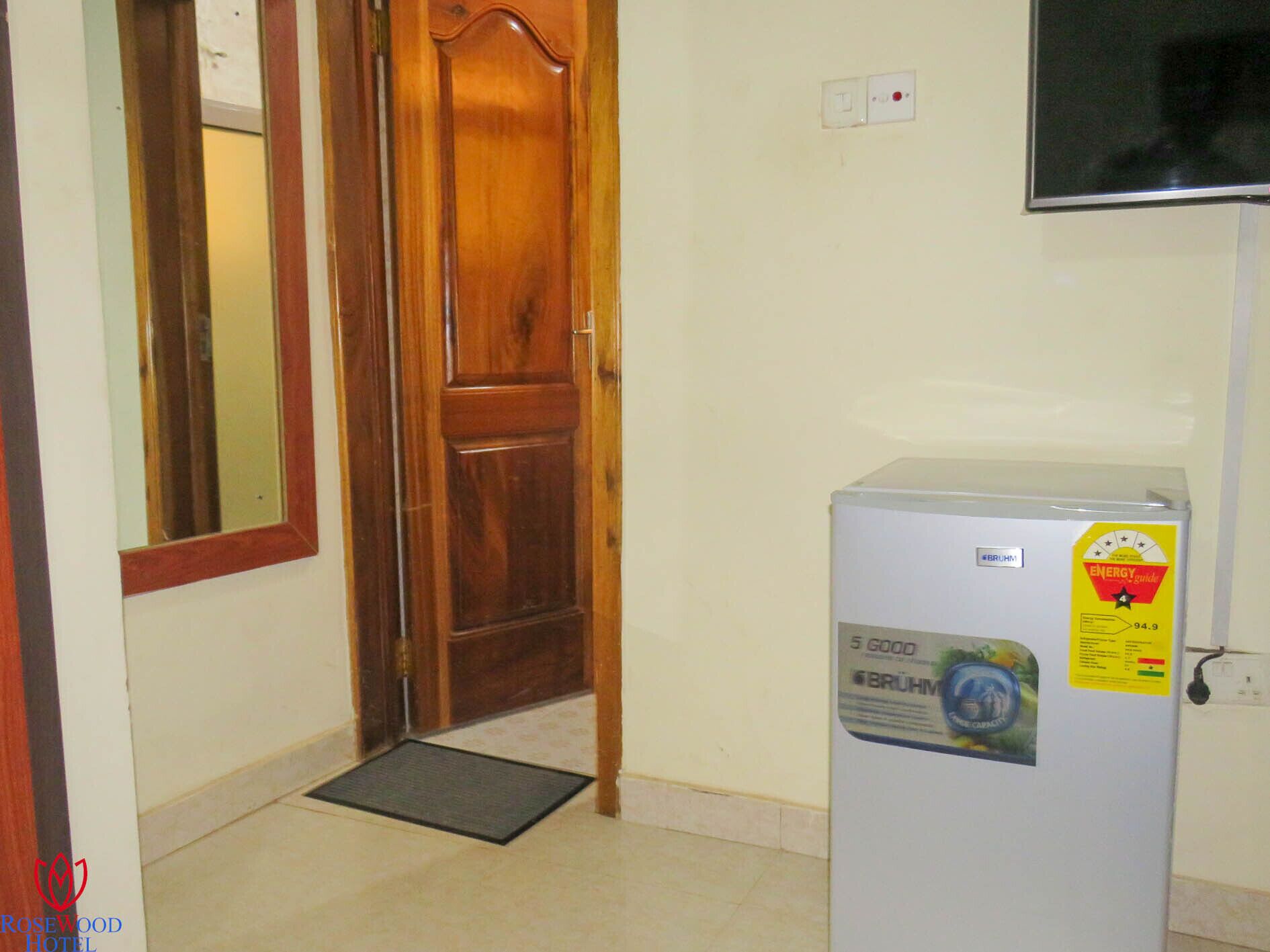 A hotel room with a wooden door, a white refrigerator with an energy efficiency sticker, a small mat at the door, and a TV mounted on the wall.