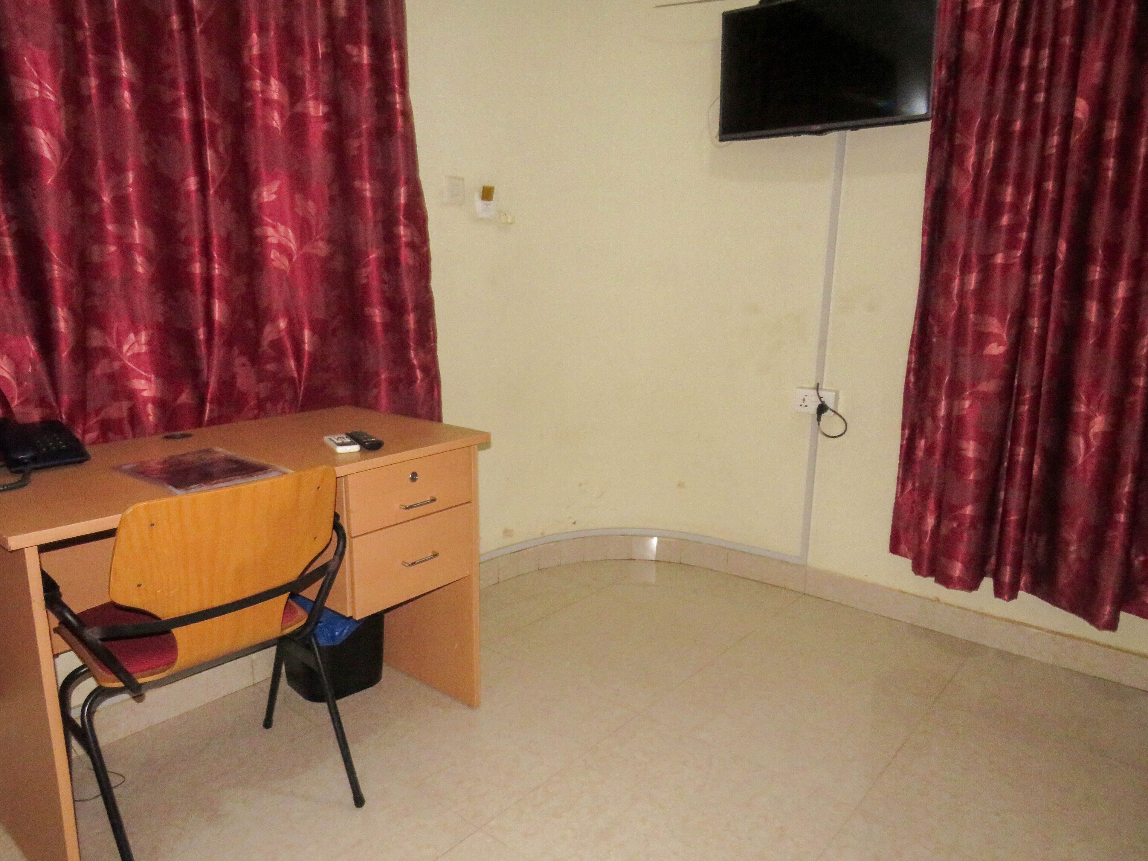 Una habitación con un escritorio de madera, una silla, un televisor montado en la pared y cortinas rojas.