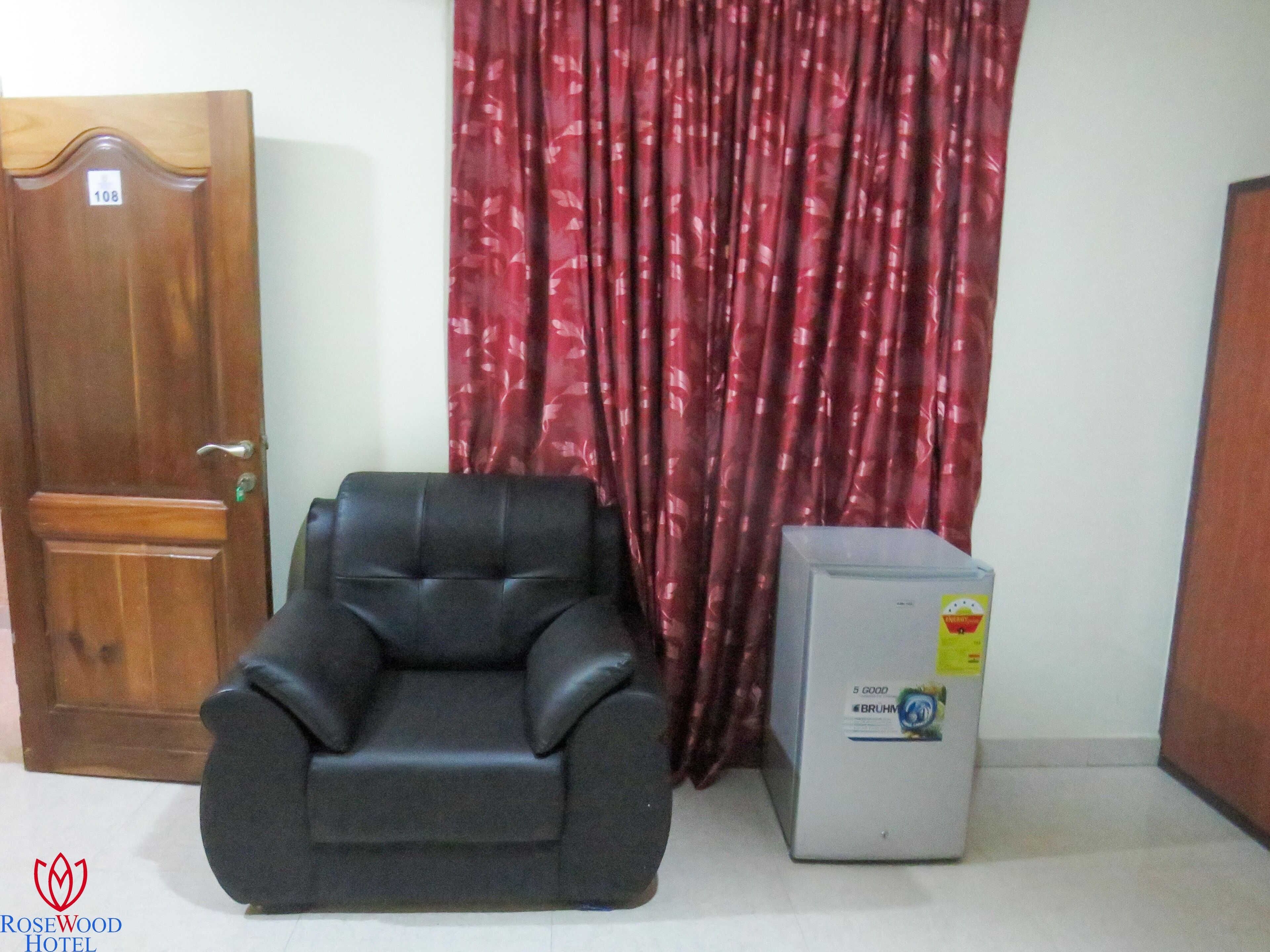 A room with a wooden door, a black leather chair, a red patterned curtain, and a white appliance with a sticker.