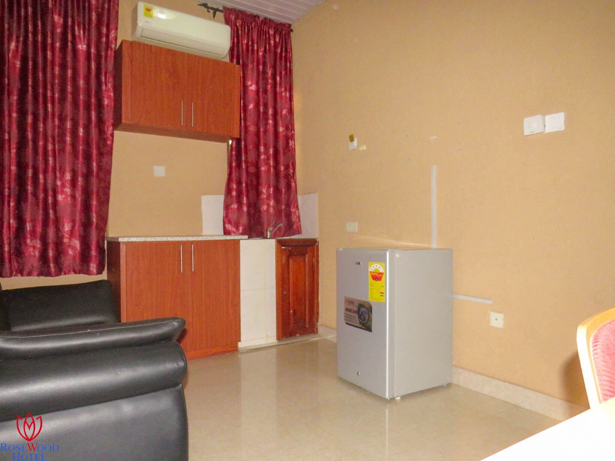 A compact hotel room with a kitchenette, a black leather chair, a small refrigerator, and a wooden cabinet.
