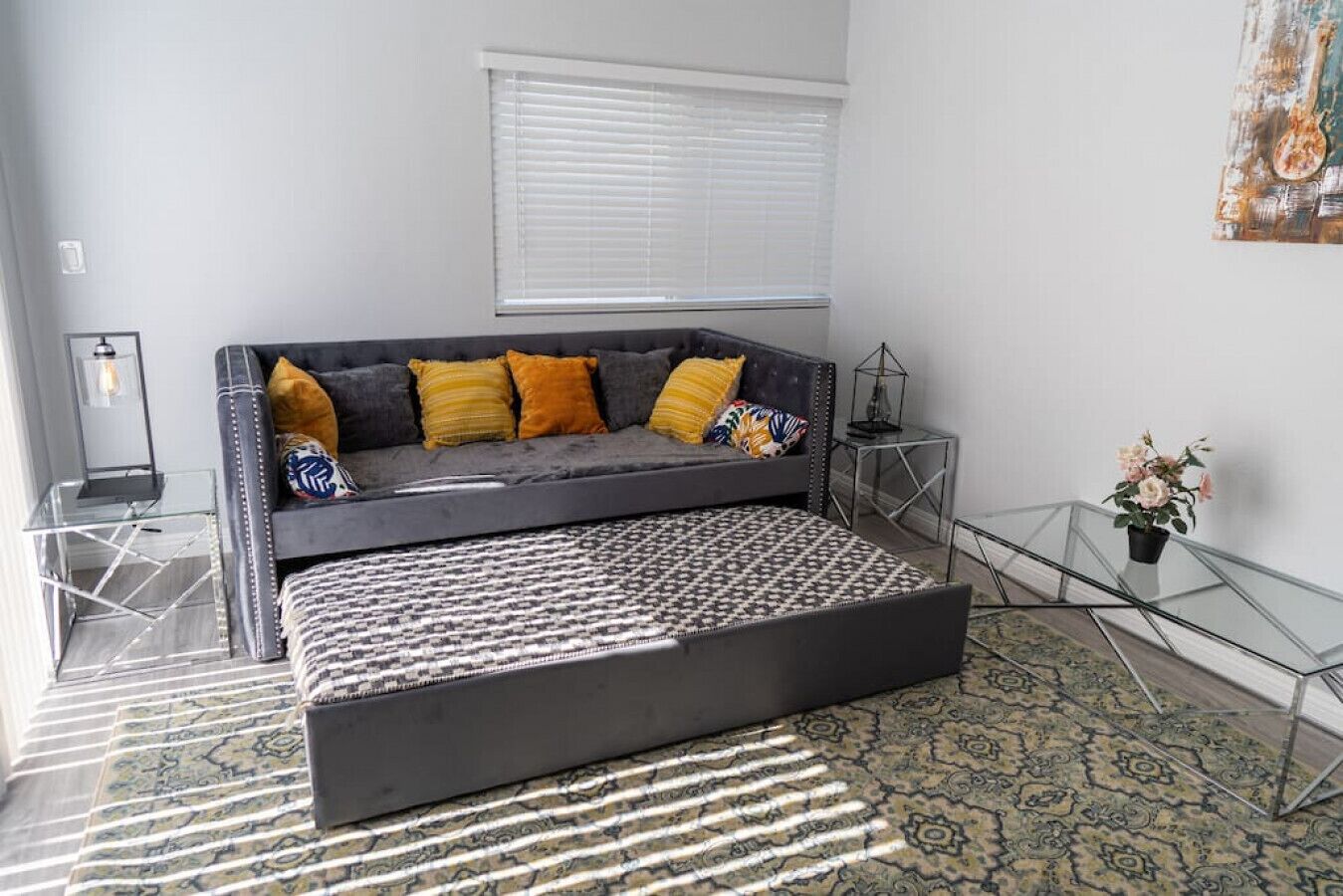 A modern living room with a grey sofa, patterned ottoman, and a glass coffee table.