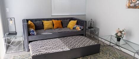 A modern living room with a grey sofa, patterned ottoman, and a glass coffee table.
