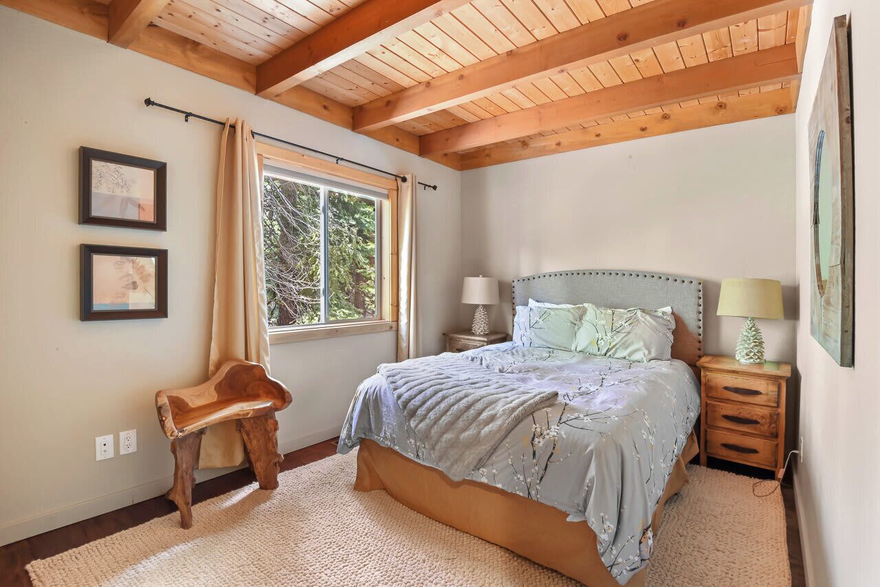 A bedroom with a wooden ceiling, a large bed with a headboard, bedside lamps, a window with curtains, and two framed pictures on the wall.