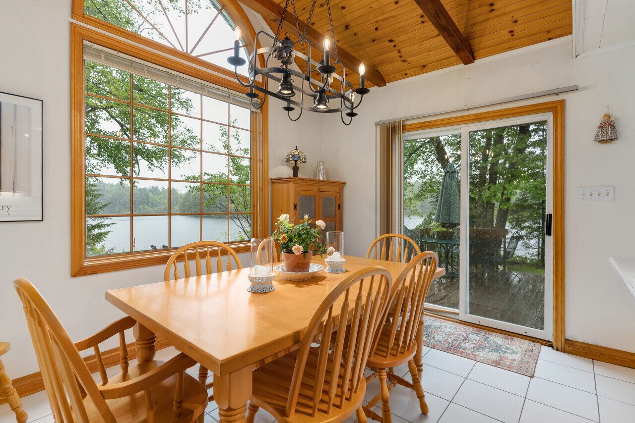 A dining area with a wooden table and chairs, large windows with a view of trees, a chandelier, and a sliding glass door leading outside.
