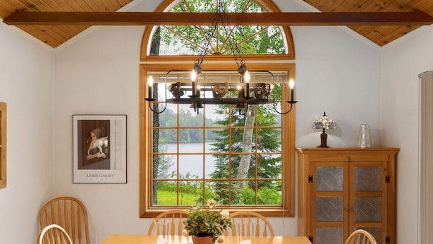 Un coin repas comprenant une table en bois, des chaises, un lustre, une grande fenêtre donnant sur des arbres et un lac, ainsi qu’un meuble avec des portes en verre.