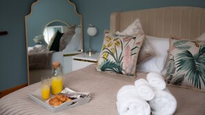 A neatly made bed with a tray of croissants, juice, and a newspaper, along with rolled towels and floral pillows.