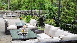An outdoor seating area with wicker furniture and a glass table, surrounded by greenery and a stone wall.