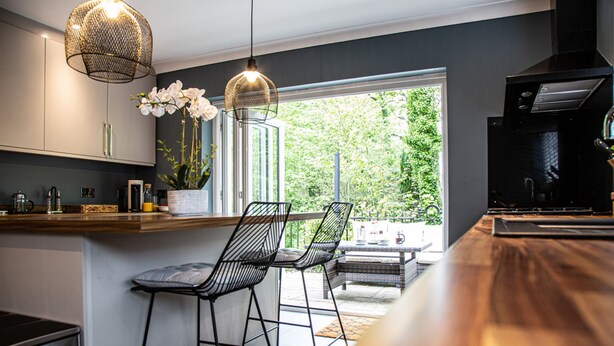 A modern kitchen with a wooden countertop, black bar stools, a built-in oven, and a large window offering a view of greenery.