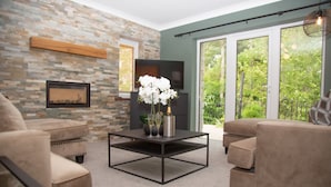 A living room with a stone wall, a flat-screen TV, a coffee table with a vase of flowers, and a large window with a view of greenery.