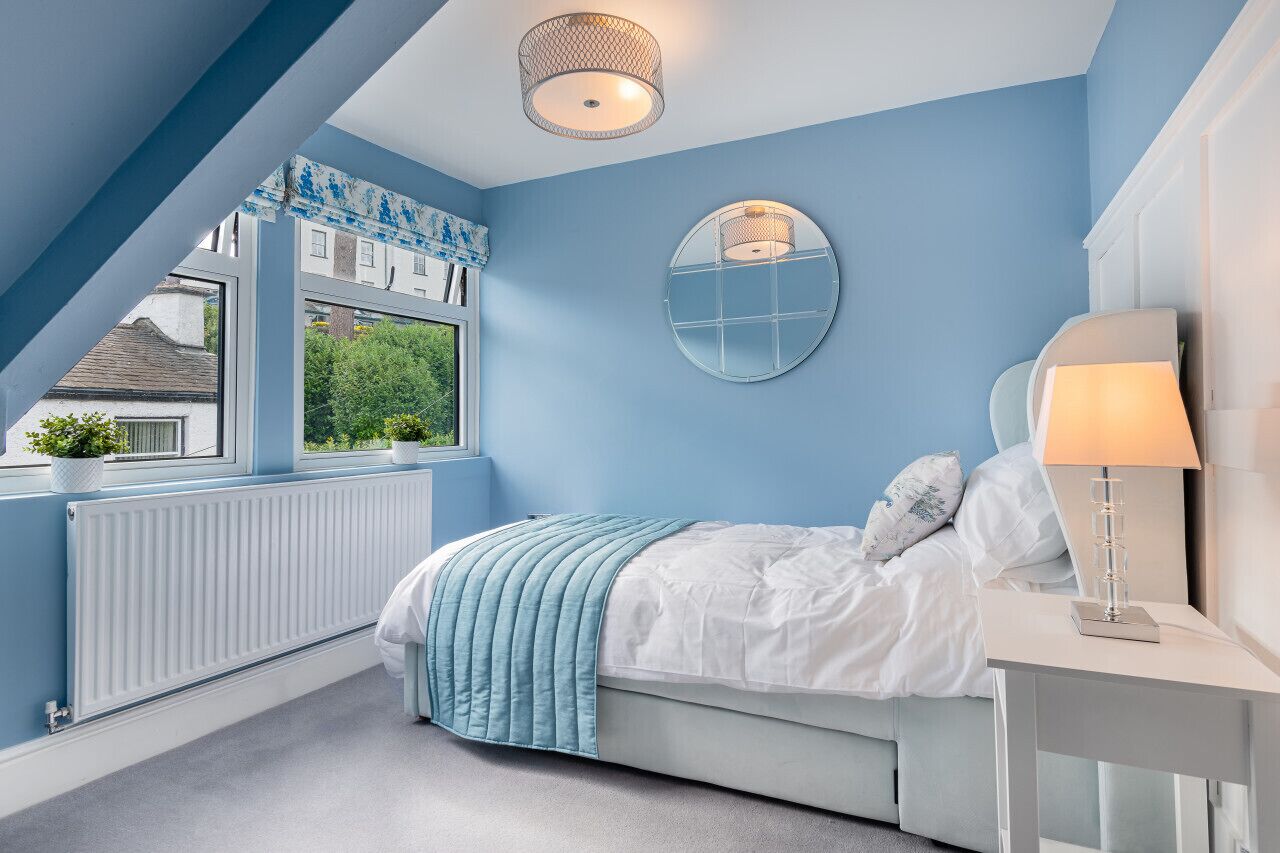 A bedroom with a sloped ceiling, a bed with a white and blue bedspread, a round mirror on the wall, and a window with a view of greenery and buildings.