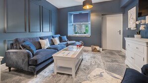 A living room with a blue sofa, a white coffee table, and a window with a view of greenery. - Belsfield Cottage in Bowness-on-windermere (Windermere)