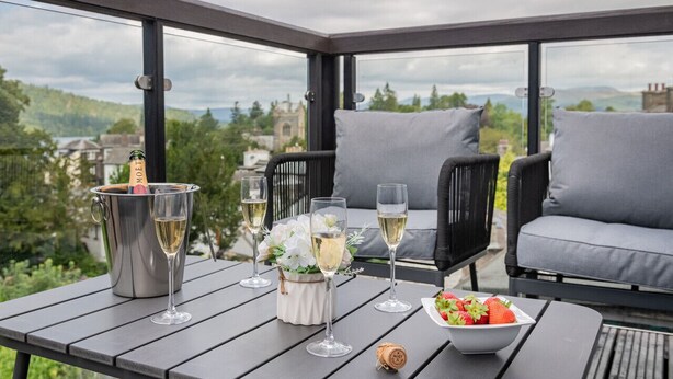 A balcony with a table set for a meal, featuring a bottle of champagne, glasses, a bowl of strawberries, and a flower arrangement. - Belsfield Cottage in Bowness-on-windermere (Windermere)