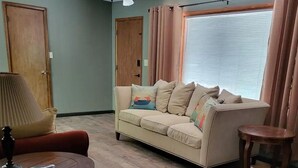Een woonkamer met een beige bank, een rode fauteuil, een klein rond tafeltje, een lamp en een raam met gordijnen.