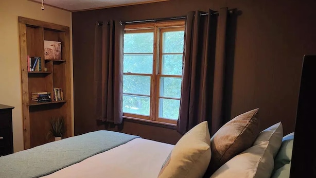 Een slaapkamer met een bed, kussens, een raam met gordijnen, een boekenplank en een bureau.