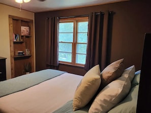 A bedroom with a bed, pillows, a window with curtains, a bookshelf, and a desk.