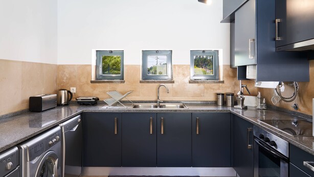 A modern kitchen with dark cabinets, a built-in washing machine, and a large window with a view of greenery. - Casa do Moinho - V3 w/ Private Pool (Abrantes)