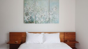 Quarto com cabeceira de madeira, roupa de cama branca e duas pinturas de flores penduradas na parede.