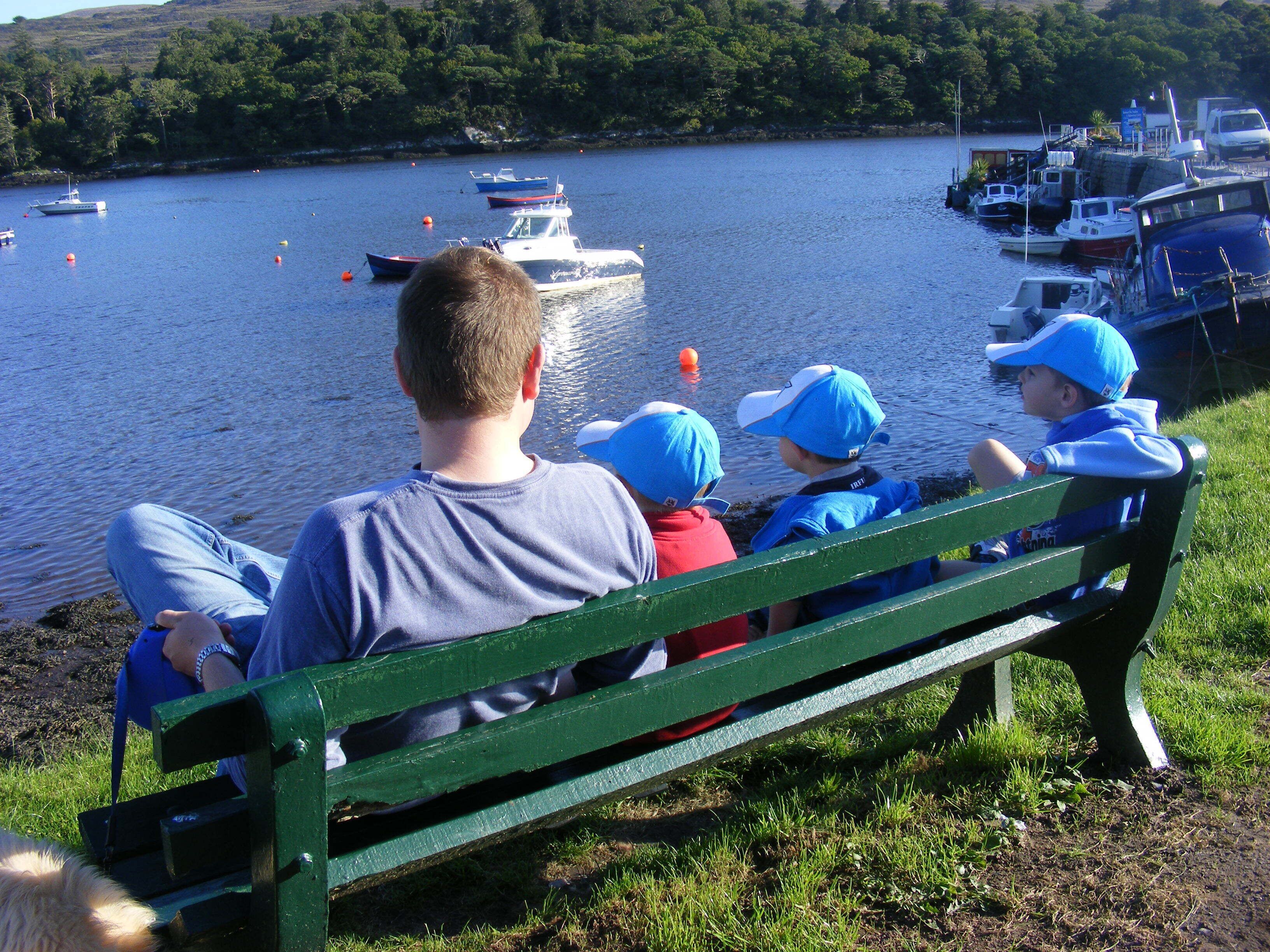 A man and two children on a green bench overlooking a lake with boats.