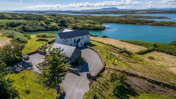 A white house with a stone driveway, surrounded by lush greenery and a body of water.