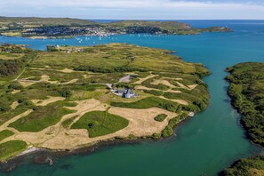 An aerial view of a coastal area with a large house surrounded by green fields and a small harbor with boats.
