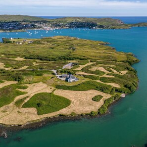An aerial view of a coastal area with a prominent house surrounded by green fields and a small harbor with boats.