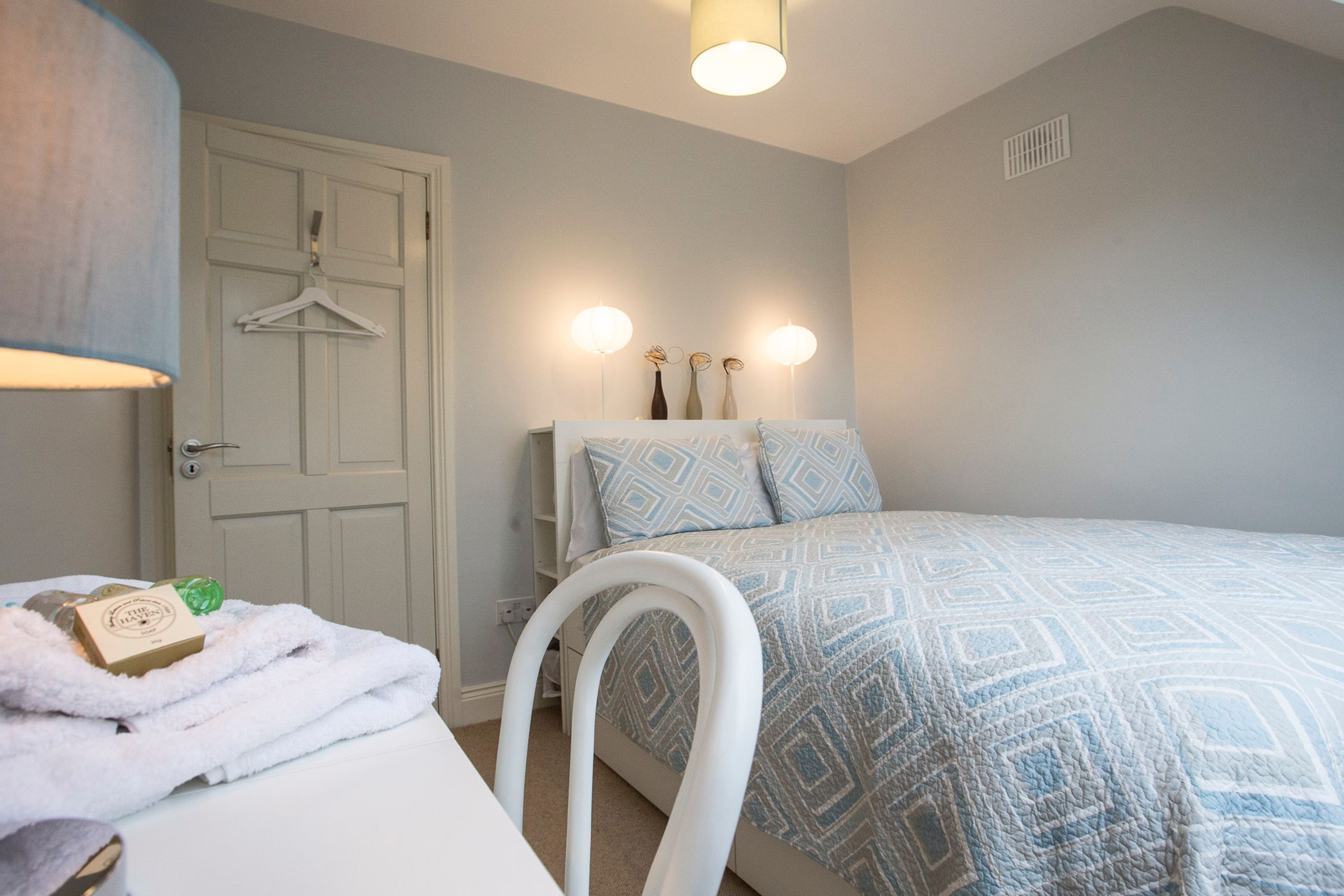 A bedroom with a bed, a chair, a hanging coat rack, and a bedside table with a towel and a bottle.