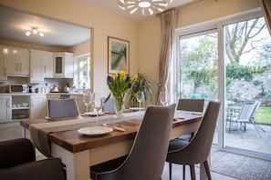 A modern dining area with a wooden table set for four, surrounded by chairs. A vase with flowers is on the table, and there is a view of a garden through the glass door. - Delightful Cottage Just Steps From Kenmare Town (Kenmare)