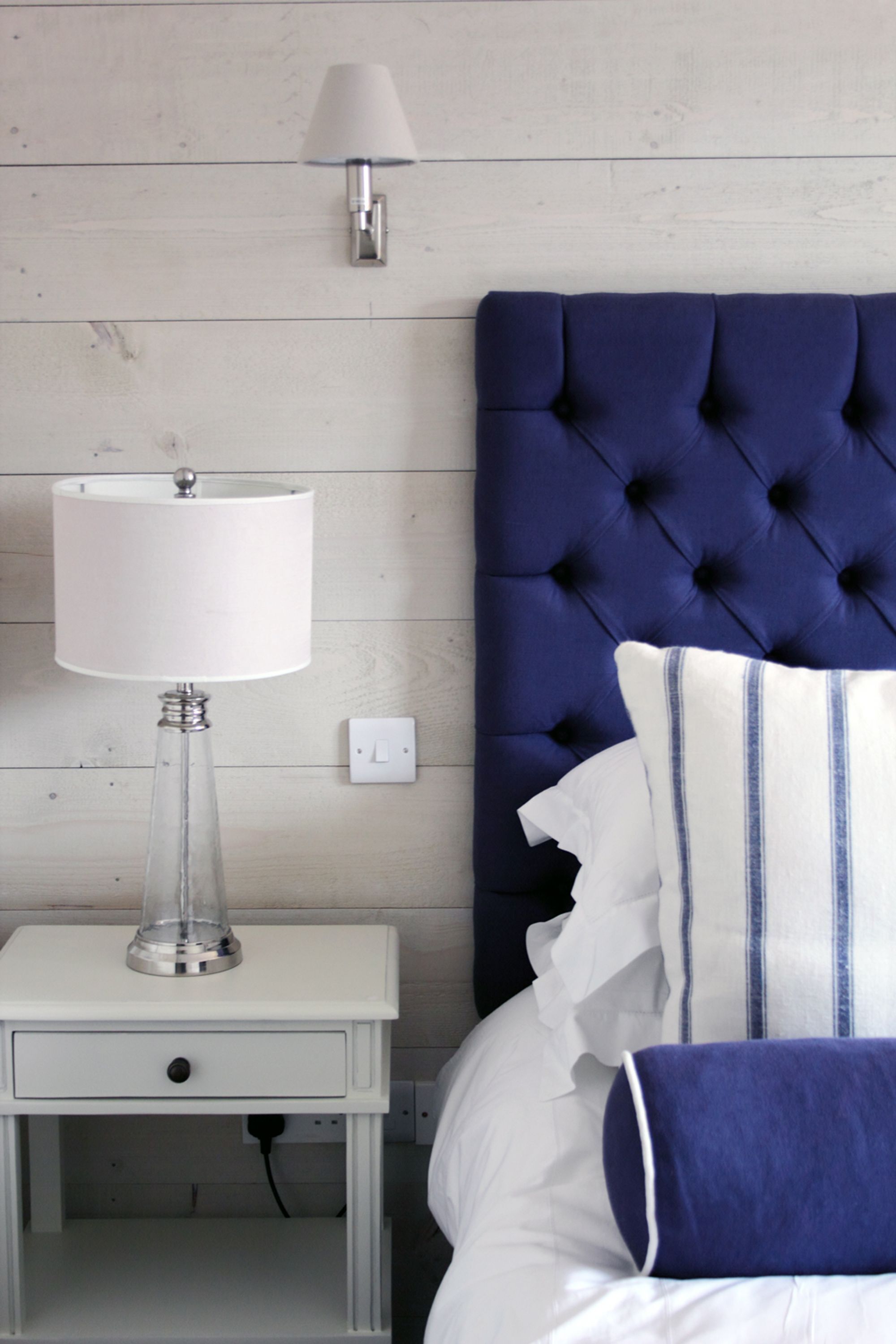 A bedroom with a white nightstand, a clear glass lamp, a white lamp, a blue tufted headboard, and white bedding with blue stripes.