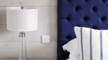 A bedroom with a white nightstand, a clear glass lamp, a white lamp, a blue tufted headboard, and white bedding with blue stripes.