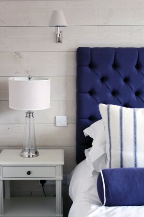 A bedroom with a white nightstand, a clear glass lamp, a white lamp, a blue tufted headboard, and white bedding with blue stripes.