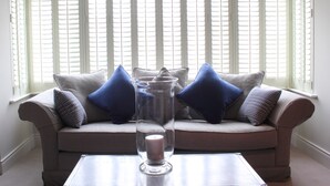 Sala de estar com sofá bege, almofadas azuis e cinzas, e um vaso de vidro transparente com uma vela sobre uma mesa de centro.