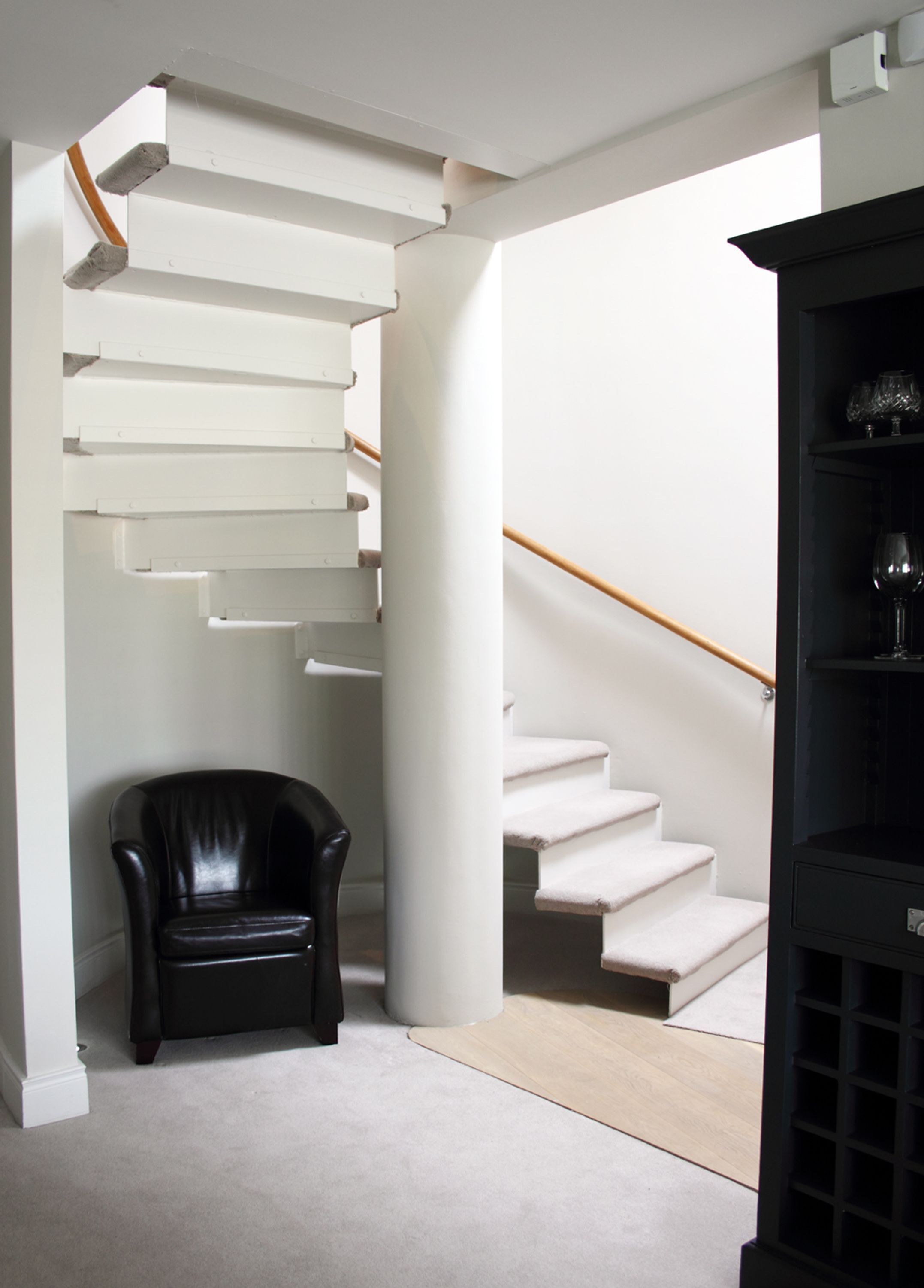 A modern staircase with white steps and a wooden handrail, leading to a black leather chair and a black shelving unit in a minimalist room.