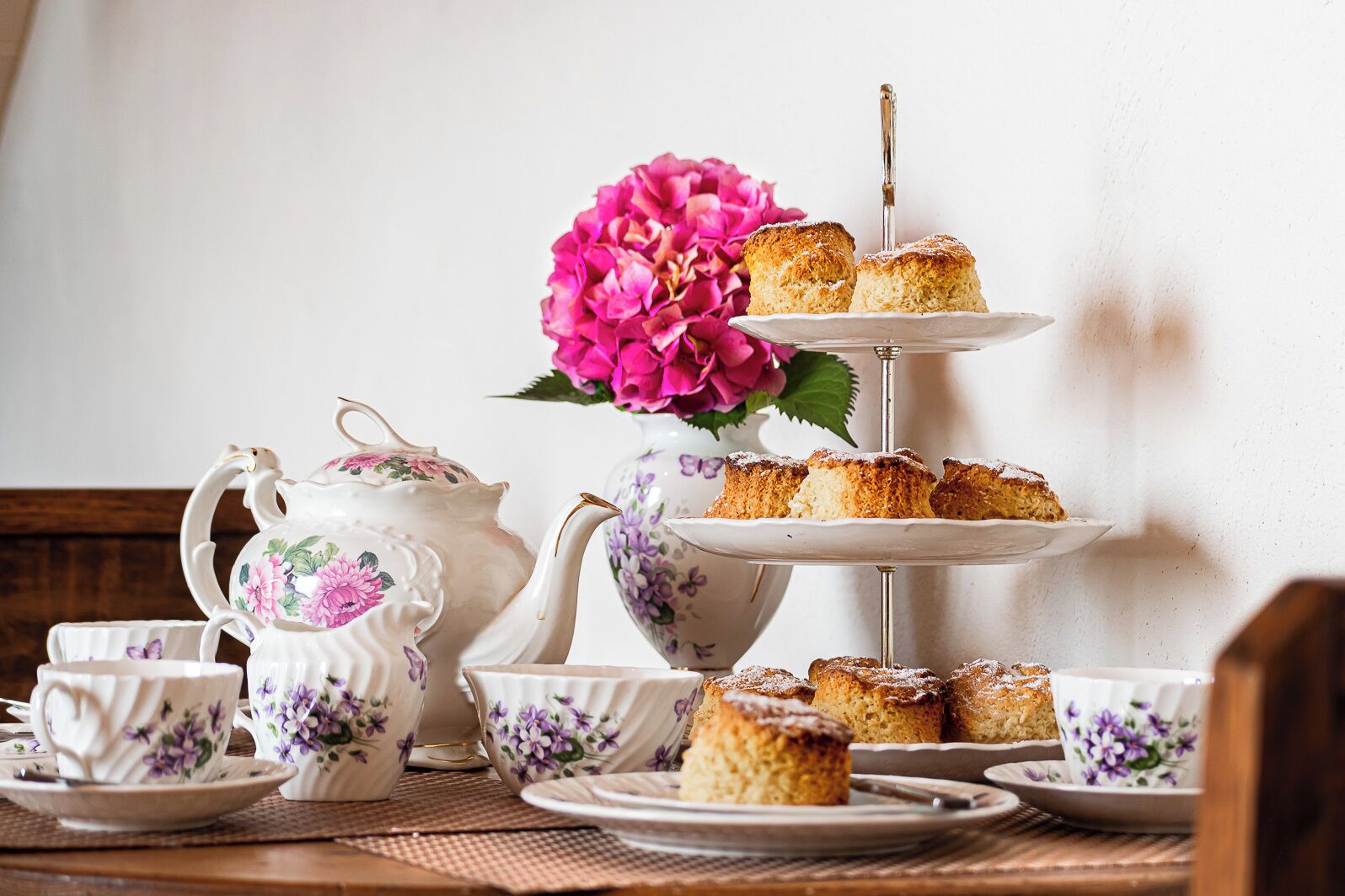 A tiered stand with pastries, a teapot, cups, and a floral arrangement on a table.