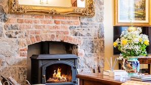 A brick fireplace with a black wood-burning stove, a wooden table with books and a vase of flowers, and a framed mirror above the fireplace. - 8 Bedroomed House Steeped in History (Ennis)